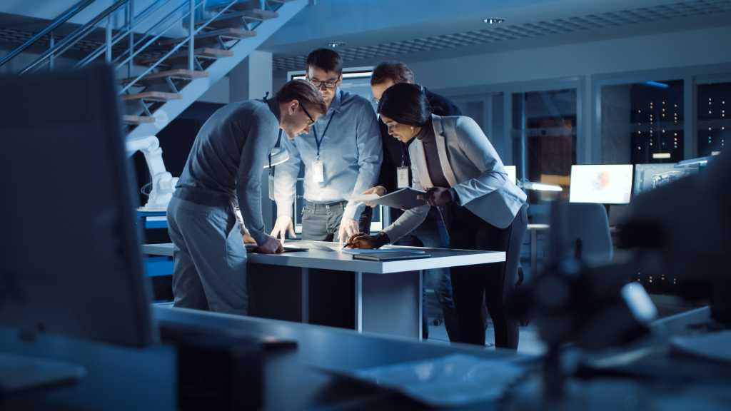 Diverse Team of Electronics Development Engineers Standing at the Desk Working with Documents, Solving Project Problems Late at Night. Specialists Working on Ultra Modern Industrial Design.