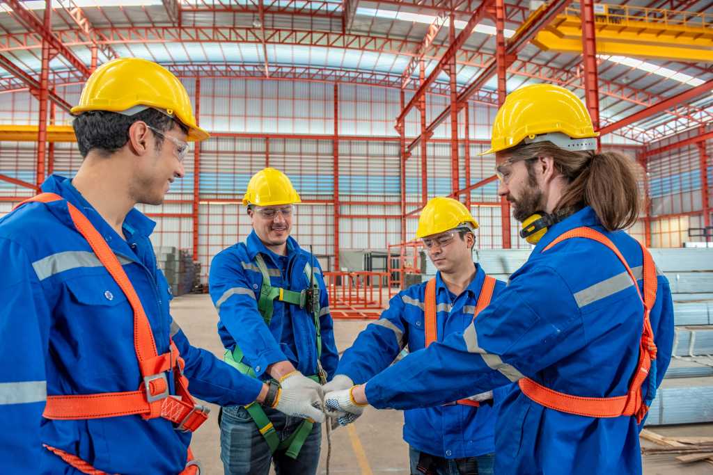 A group of construction workers wearing hard hats and safety gear standing in a circle with their hands together.